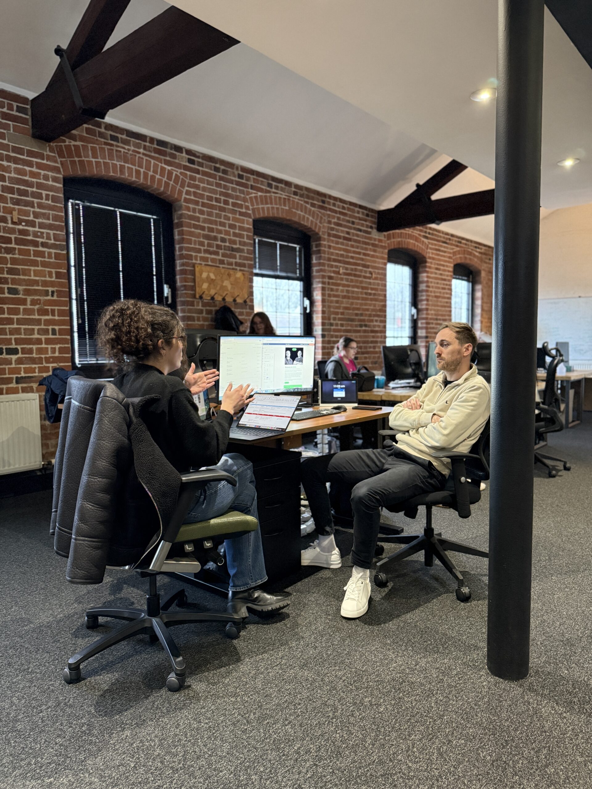 Two team members having a discussion at a desk in a rustic brick-walled office, with colleagues working in the background