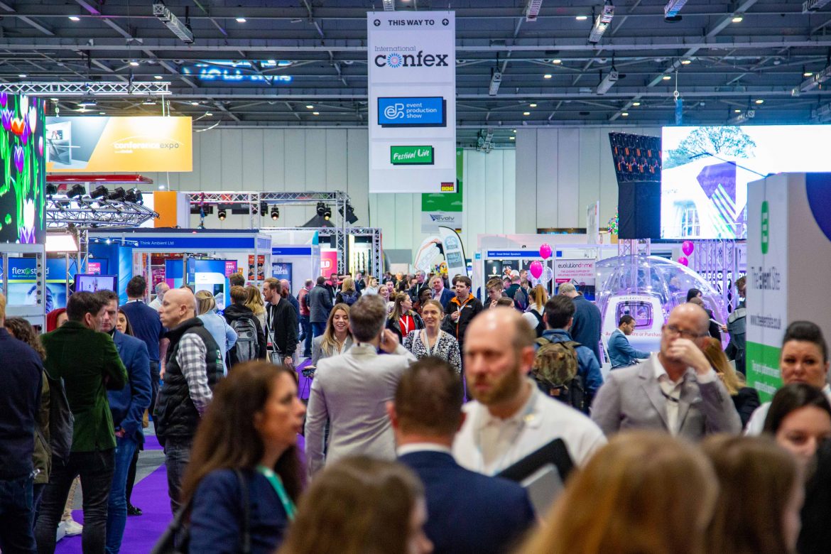 An indoor trade show setting at International Confex, featuring overhead signage pointing the way to the event, large booth structures with graphic panels, and a busy exhibition space under a high-ceiling venue.